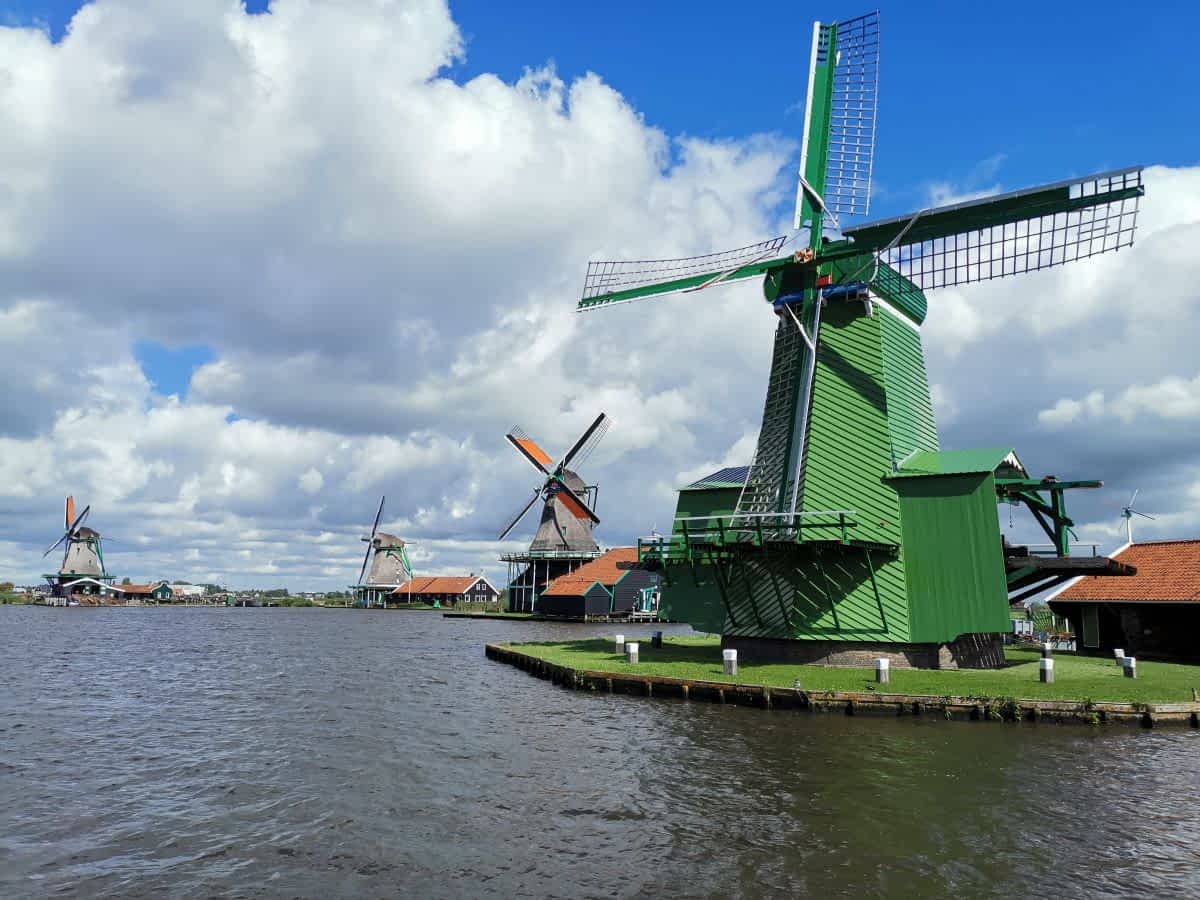 Blick über eine Gracht auf mehrere historische Windmühlen an der Zaanse Schans unter einem bewölkten Himmel. Im Vordergrund eine auffällig grüne Windmühle. Blick über eine Gracht auf mehrere historische Windmühlen an der Zaanse Schans unter einem bewölkten Himmel. Im Vordergrund eine auffällig grüne Windmühle.