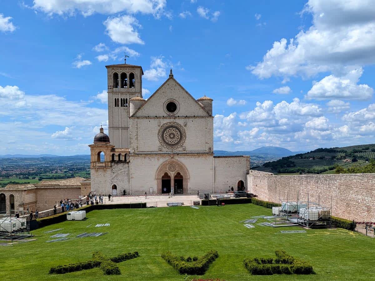 Die Basilika San Francesco in Assisi mit der prächtigen Fassade und dem Glockenturm, umgeben von gepflegten Grünflächen unter blauem Himmel.