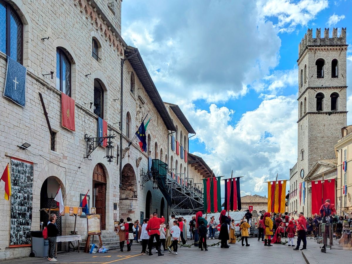 Belebter Platz in Assisi mit historischen Gebäuden und festlich gekleideten Menschen während des Calendimaggio Festes. Die Piazza del Comune ist geschmückt mit Fahnen und Wappen, im Hintergrund ist der Torre del Popolo zu sehen.