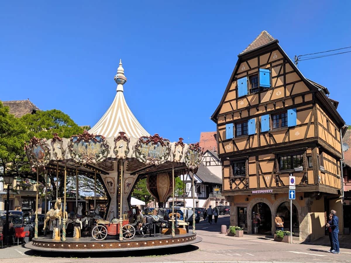 Ein nostalgisches, weiß-gestreiftes Karussell auf einem öffentlichen Platz in Obernai neben einem hohen Fachwerkhaus mit blauen Fensterläden.