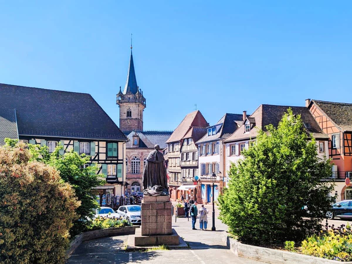 Marktplatz in Obernai im Elsass mit historischen Fachwerkhäusern, einem Denkmal im Vordergrund und dem markanten Kappelturm unter blauem Himmel.