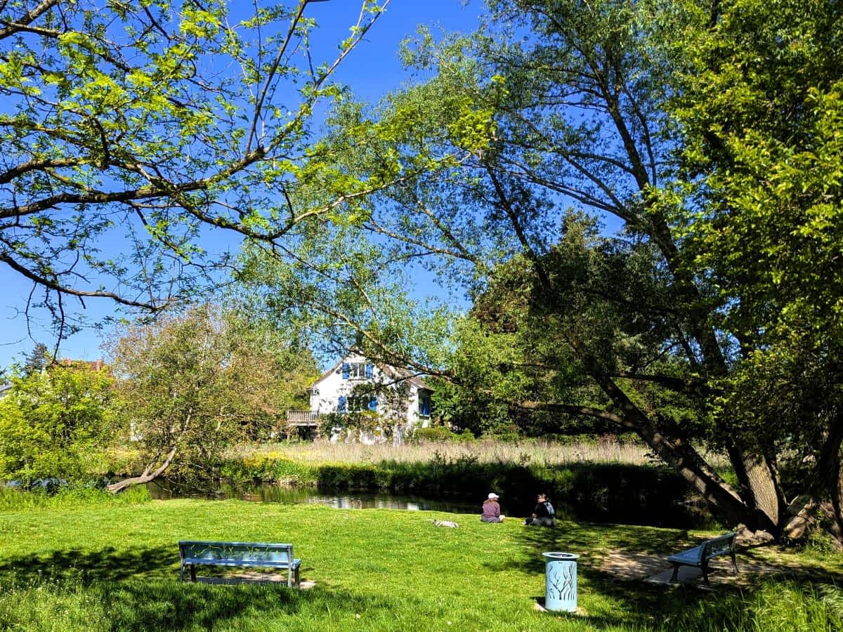 in idyllischer Rastplatz im Grünen an einem Flussufer mit Parkbänken und einem weißen Haus im Hintergrund.