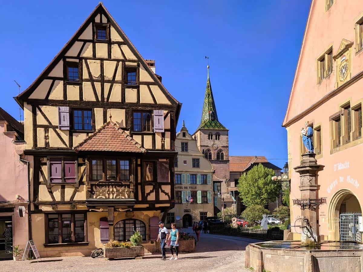 Historischer Marktplatz in Turckheim mit einem großen, verzierten Fachwerkhaus, einem Brunnen im Vordergrund und dem bunt gedeckten Kirchturm unter blauem Himmel.