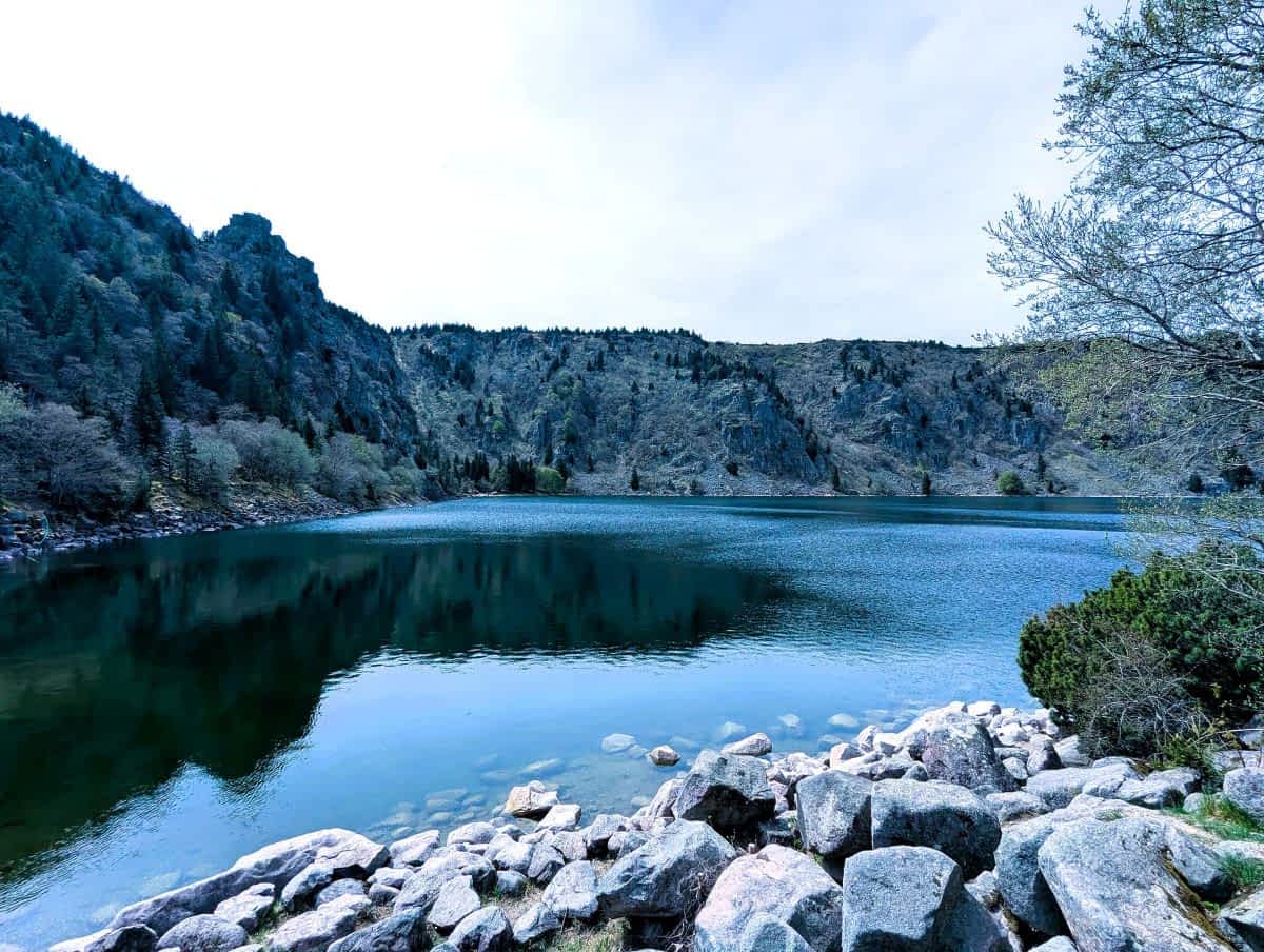 Ein klarer, blauer Bergsee (Lac Blanc) in den Vogesen, umgeben von steilen, bewaldeten Felswänden und grauen Granitblöcken im Vordergrund