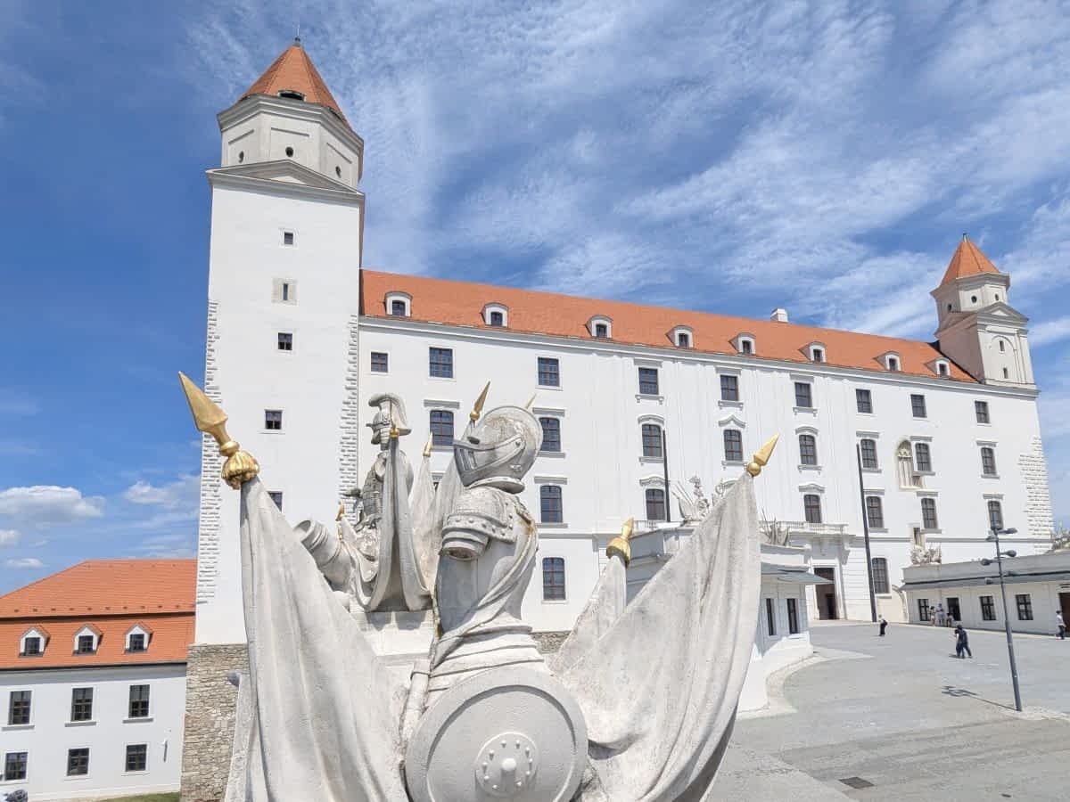 Nahaufnahme der hellen, restaurierten Fassade der Bratislavaer Burg mit ihren Ecktürmen. Im Vordergrund, auf der Brüstung, eine steinerne Trophäenskulptur mit antiken Rüstungen und Flaggen