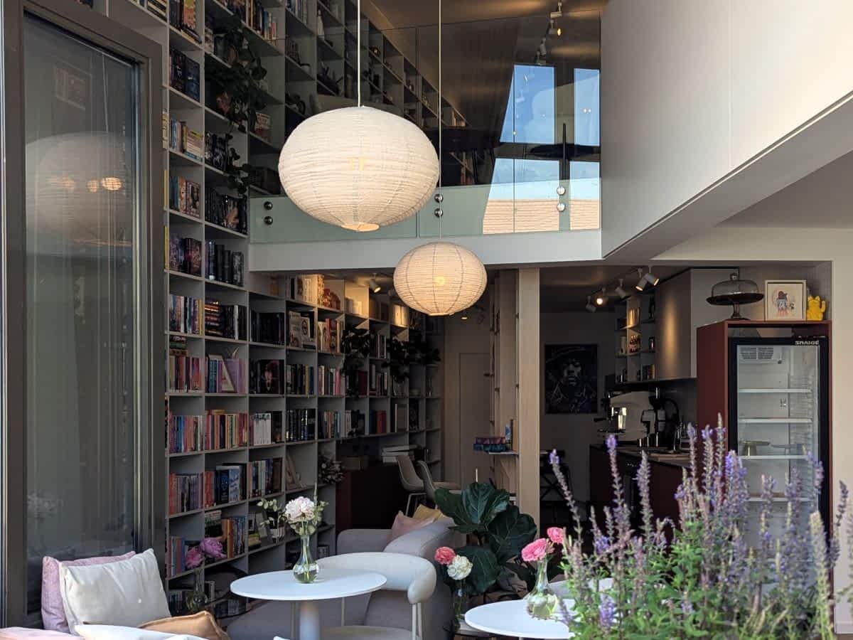Gemütlicher Innenraum eines Cafés in Bratislava mit einer hohen, bis zur Decke reichenden Bücherwand. Zwei weiße kugelförmige Lampen hängen von der Decke über kleinen Tischen mit frischen Blumen, im Hintergrund ist eine Bar und ein Kühlschrank erkennbar.