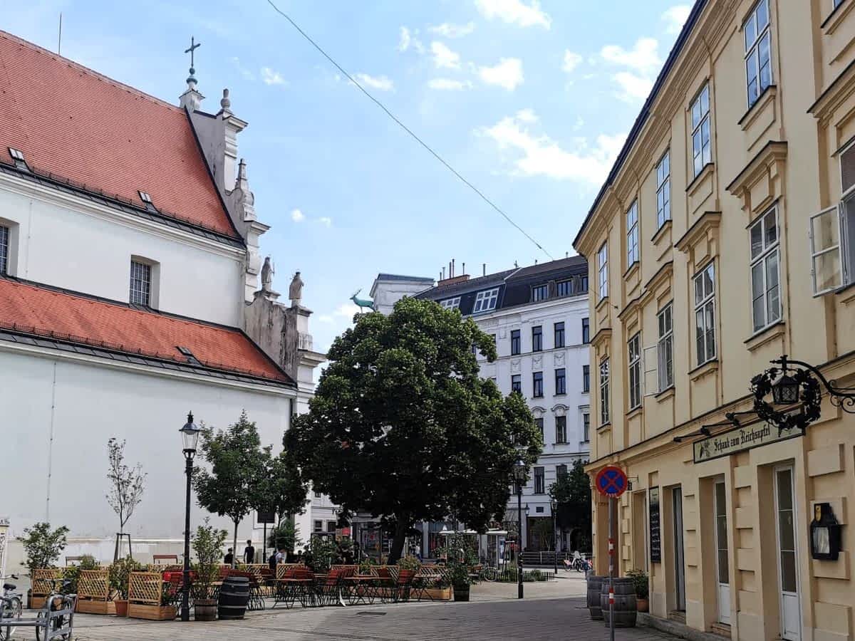 Ansicht des Karmeliterplatzes in Wien mit der barocken Karmeliterkirche auf der linken Seite und der Gastterrasse 'Schenke zum Reichsapfel' auf der rechten Seite, umgeben von Bäumen unter blauem Himmel.
